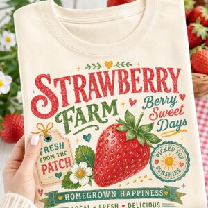 May include: A cream-colored t-shirt featuring a graphic print with the text "STRAWBERRY FARM," "Berry Sweet Days," "FRESH FROM THE PATCH," "PICKED FOR SUNSHINE," and "HOMEGROWN HAPPINESS." The design includes a large red strawberry, a white flower, and small hearts.