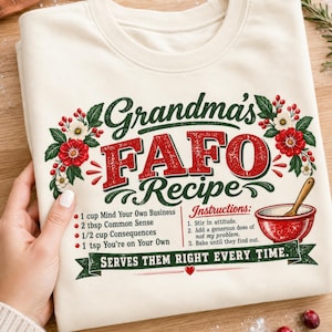 May include: A cream-coloured sweatshirt featuring a graphic with the text Grandma's FAFO Recipe. The design includes red and green floral accents, a red mixing bowl with a wooden spoon, and a list of ingredients and instructions in black text.