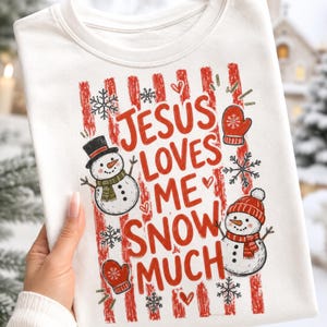 May include: A white crewneck sweatshirt featuring a red and white striped background graphic with the text JESUS LOVES ME SNOW MUCH. The design includes two snowmen, one wearing a black top hat and one in a red beanie, plus red mittens and snowflakes.