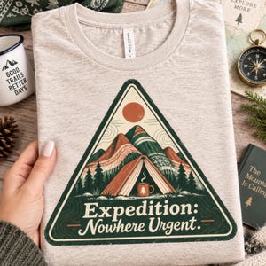 Op de afbeelding: Een beige gemêleerd t-shirt met een driehoekige afbeelding van een tent tussen bergen en dennenbomen onder een zon. Het ontwerp bevat een dampende mok en de tekst "Expedition: Nowhere Urgent." Het t-shirt heeft een ronde halslijn.