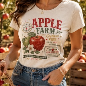 May include: A cream-colored short-sleeve t-shirt featuring a graphic print with the text APPLE FARM, Orchard Cutie, Crisp Cozy Charm, Fresh From Our Orchard, Hand Picked With Care, Sweet Memories Local Goodness, and Est. 1987, alongside red and green apples.