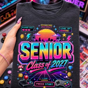 Può includere: T-shirt grigio scuro con un colorato design retrò a tema videogioco. Il design include le parole "SENIOR Class of 2027" in un font pixelato, insieme a elementi grafici a tema gioco come un joystick, un controller e un tramonto.