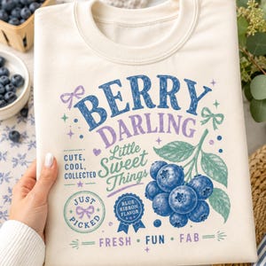 May include: A cream-colored crewneck sweatshirt featuring a graphic print of blueberries on a branch with the text BERRY DARLING, Little Sweet Things, CUTE, COOL, COLLECTED, JUST PICKED, BLUE RIBBON FLAVOR, and FRESH, FUN, FAB with small bow icons.