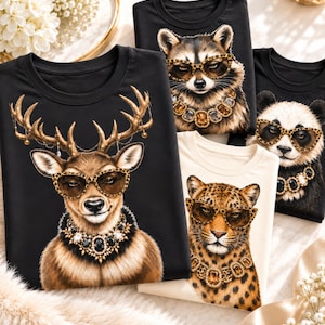 Pode incluir: Uma coleção de t-shirts com estampas gráficas de um veado, um guaxinim, um panda e um leopardo. Cada animal usa óculos com estampa de leopardo e um grande colar ornamentado com joias. As t-shirts são pretas ou de cor creme.