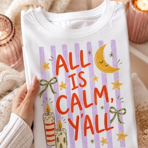 May include: A white t-shirt featuring a graphic print with vertical light purple stripes and the red text "ALL IS CALM, Y'ALL". The design includes a yellow crescent moon with a face, yellow stars, two green bows, and two lit candles.