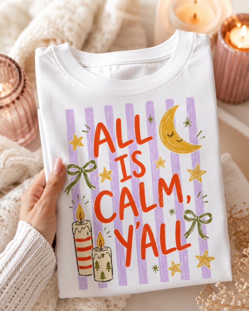 May include: A white t-shirt featuring a graphic print with vertical light purple stripes and the red text "ALL IS CALM, Y'ALL". The design includes a yellow crescent moon with a face, yellow stars, two green bows, and two lit candles.