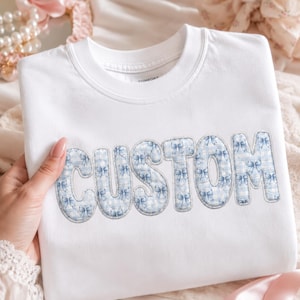 May include: White sweatshirt with the word "CUSTOM" embroidered in a blue bow pattern. The sweatshirt is a crew neck style and appears to be made of a soft material. The background includes pearls and pink ribbons.