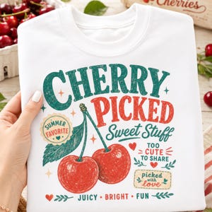 May include: A white crewneck sweatshirt featuring a graphic print of two red cherries with green stems and leaves. The text reads CHERRY PICKED Sweet Stuff, SUMMER FAVORITE, TOO CUTE TO SHARE, picked with love, and JUICY BRIGHT FUN in red and green.