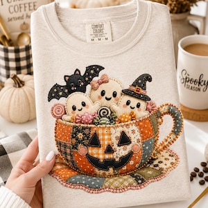 May include: A light beige crewneck sweatshirt featuring a graphic of a patchwork pumpkin-shaped teacup filled with three small ghosts, a black bat, and colorful candies. The teacup sits on a matching patchwork saucer. The inner label reads Comfort Colors.
