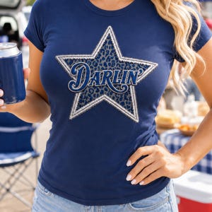 May include: Navy blue t-shirt featuring a star design with a leopard print center and the word "Darlin" in a script font. The star is outlined in white and silver. The shirt is paired with denim shorts.