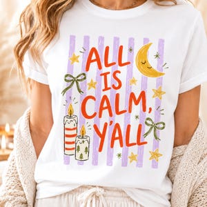 May include: A white short-sleeved t-shirt featuring a graphic print with vertical lavender stripes. The text reads ALL IS CALM, Y'ALL in red letters, surrounded by illustrations of a yellow crescent moon, gold stars, two lit candles, and green bows.