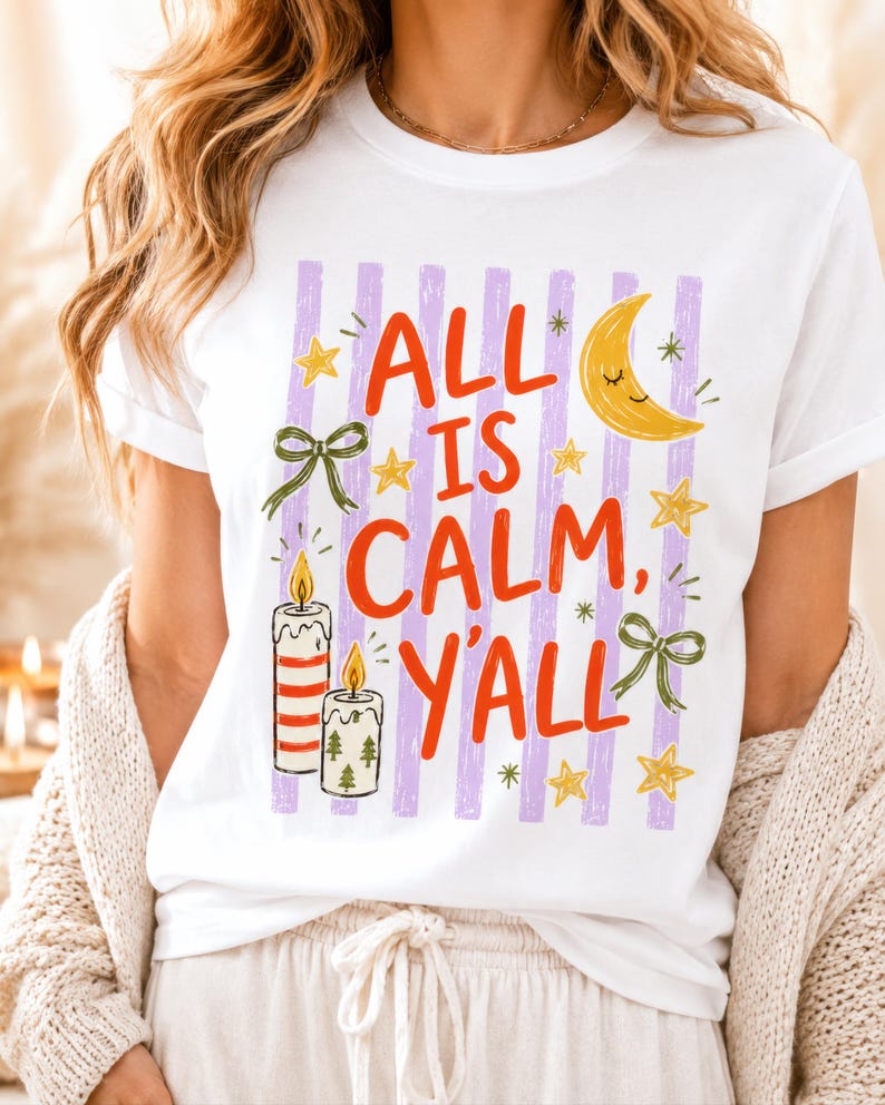 May include: A white short-sleeved t-shirt featuring a graphic print with vertical lavender stripes. The text reads ALL IS CALM, Y'ALL in red letters, surrounded by illustrations of a yellow crescent moon, gold stars, two lit candles, and green bows.