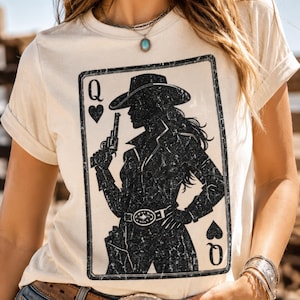 May include: Cream-colored t-shirt featuring a playing card design with a silhouette of a cowgirl holding a gun. The card is the Queen of Hearts and Spades. The shirt has short sleeves and a crew neck.