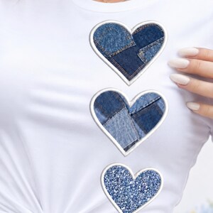 May include: White t-shirt with three heart-shaped patches. The top two hearts are made of denim patches, and the bottom heart is covered in blue glitter. Each heart has a white border.