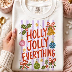 May include: A white crewneck sweatshirt featuring a graphic print with the text "HOLLY JOLLY EVERYTHING" in red letters. The design includes purple vertical stripes, green holly leaves, red berries, gold bells, and colorful Christmas ornaments.