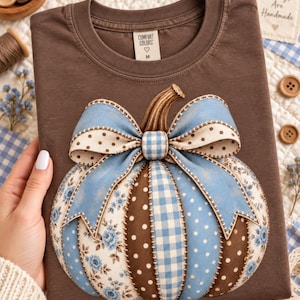 Op de afbeelding: Een donkerbruin t-shirt met een grote pompoenafbeelding. De pompoen bestaat uit afwisselende panelen met blauwe bloemen, bruine stippen en blauwe ruitjes, bekroond met een grote blauwe strik met crèmekleurige stippen en een bruine steel.