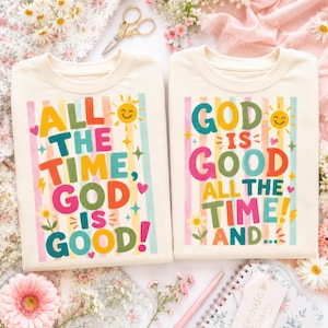 Puede incluir: Dos camisetas de cuello redondo color crema con rayas verticales coloridas y texto gráfico. La camiseta de la izquierda dice "ALL THE TIME, GOD IS GOOD!" y la de la derecha "GOD IS GOOD ALL THE TIME AND..." con iconos de sol, estrellas, corazones y flores.