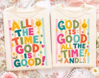 May include: Two cream-colored crewneck t-shirts featuring colorful vertical stripes and graphic text. The left shirt reads "ALL THE TIME, GOD IS GOOD!" and the right shirt reads "GOD IS GOOD ALL THE TIME AND..." with sun, star, heart, and flower icons.