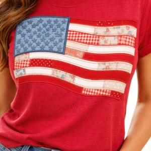 May include: Red t-shirt with a patchwork American flag design. The flag features a blue field with white stars and red and white stripes made of patterned fabrics. The shirt has short sleeves and a crew neck.