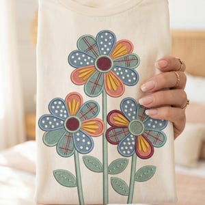 Puede incluir: Camiseta color crema con un diseño floral bordado. El diseño incluye tres flores con pétalos azules, amarillos y rojos, tallos y hojas verdes. Las flores tienen un patrón de patchwork con lunares y cuadros.