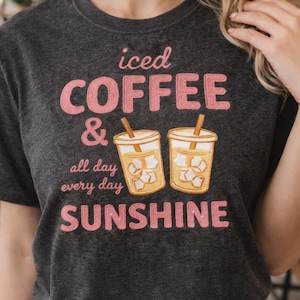 Puede incluir: Camiseta gris oscuro con el texto "iced COFFEE & all day every day SUNSHINE" en rosa. La camiseta presenta dos tazas de café helado bordadas con cubitos de hielo y pajitas.