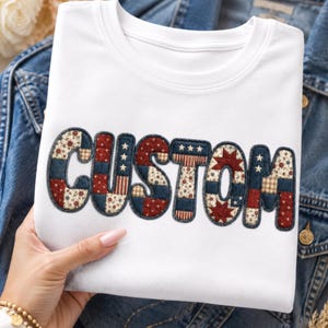 May include: White crew neck t-shirt with the word "CUSTOM" in a patchwork design. The letters are in red, navy, and cream fabrics with star and floral patterns. The shirt is folded on a denim jacket.