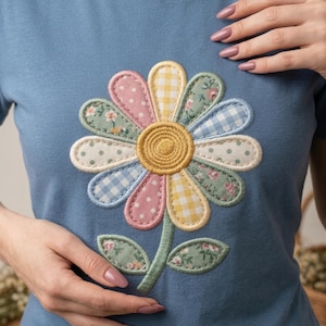 Puede incluir: Camiseta azul claro con una gran margarita bordada. Los pétalos de la margarita presentan varios estampados pastel, incluyendo lunares, cuadros vichy y estampados florales. El centro es amarillo, y el tallo y las hojas son verdes.