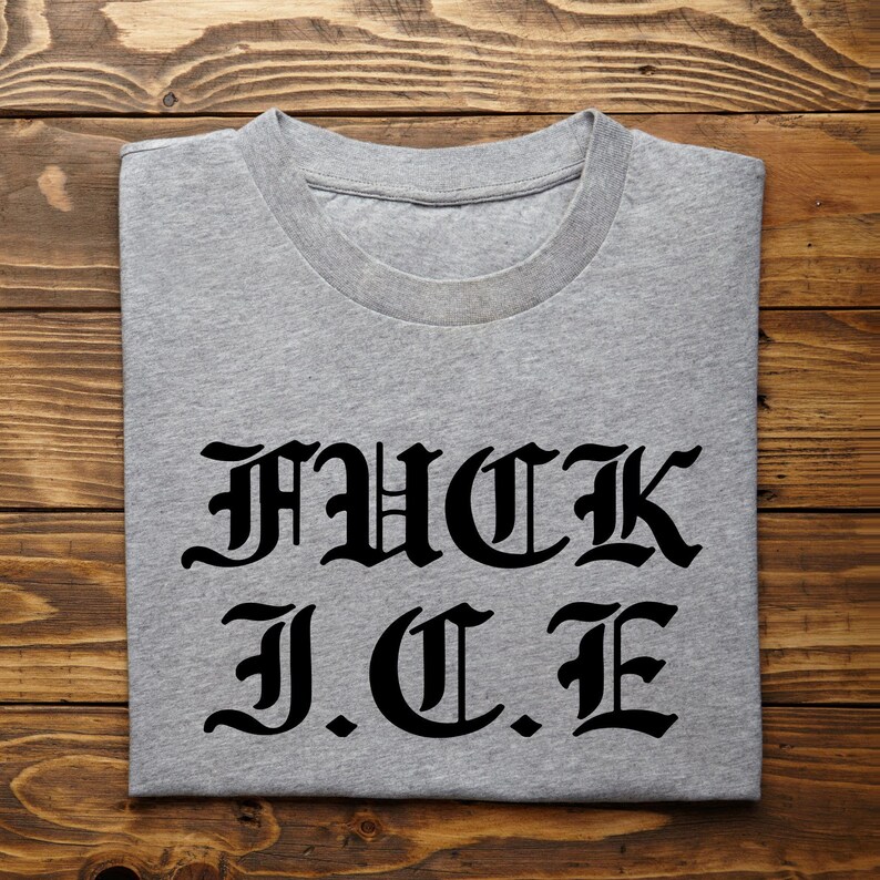 May include: A grey t-shirt featuring the text "FUCK I.C.E" in a gothic font. The folded shirt is displayed on a wooden surface.