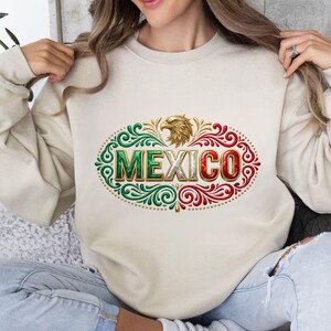 May include: A beige crewneck sweatshirt featuring a central graphic of a golden eagle head above the word MEXICO in block letters. The text and surrounding ornate filigree patterns are colored in shades of green, white, and red.