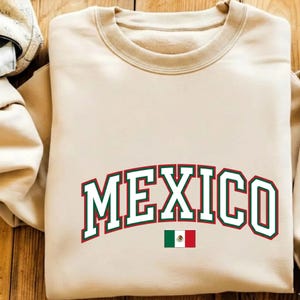 May include: A beige crewneck sweatshirt featuring the word MEXICO printed in a large, arched, collegiate-style font with white lettering and dark green and red outlines. Below the text is a small, rectangular flag of Mexico.
