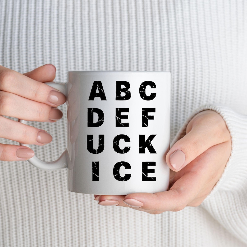 May include: White ceramic mug with black text spelling out "ABC DEF UCK ICE". The mug is held by a person wearing a white jumper. The letters have a distressed, textured appearance.