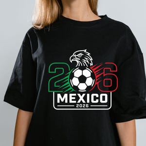 May include: A black t-shirt featuring a graphic design with the number 2026, a soccer ball, and an eagle head. The number 2 is green, the number 6 is red, and the text MEXICO 2026 is printed in white within a rectangular border on the chest.