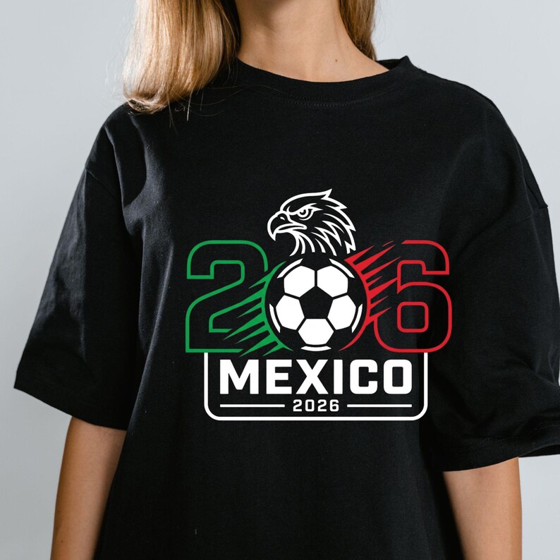May include: A black t-shirt featuring a graphic design with the number 2026, a soccer ball, and an eagle head. The number 2 is green, the number 6 is red, and the text MEXICO 2026 is printed in white within a rectangular border on the chest.