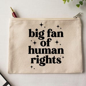 May include: A beige canvas zippered pouch with the text "big fan of human rights" in black, with star accents. A pen is visible in the open zipper. The pouch is rectangular and appears to be made of a sturdy fabric.