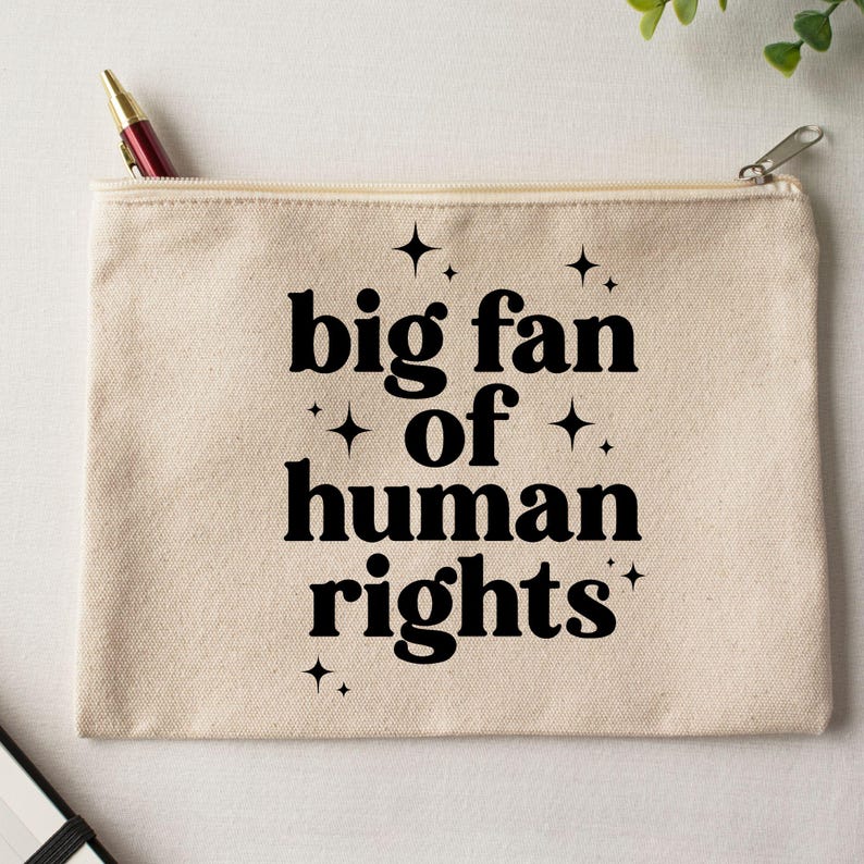 May include: A beige canvas zippered pouch with the text "big fan of human rights" in black, with star accents. A pen is visible in the open zipper. The pouch is rectangular and appears to be made of a sturdy fabric.