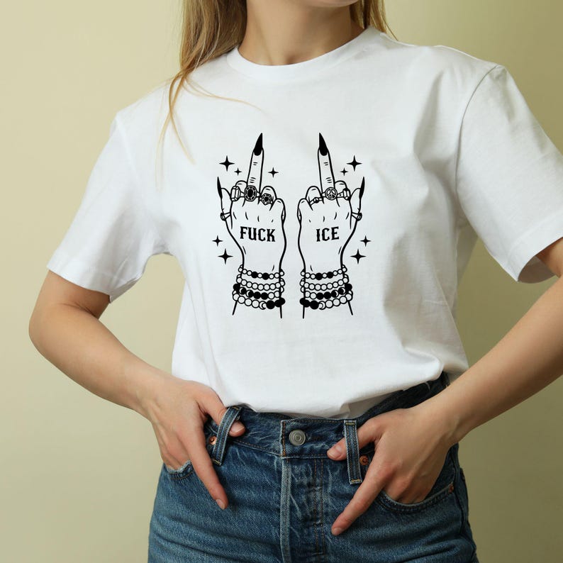 May include: White t-shirt with a black graphic of two hands displaying the middle finger. The hands are decorated with rings and bracelets, and the text "FUCK ICE" is shown. The design includes star accents.
