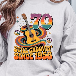 May include: White sweatshirt with a retro design featuring a guitar, peace signs, and the text "70 Still Groovin' Since 1956" in orange and yellow. The design has a groovy, vintage aesthetic.