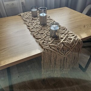 Puede incluir: Camino de mesa de macramé beige con diseño de rombos y flecos. Tres candelabros de vidrio con velas azules están colocados sobre el camino de mesa, sobre una mesa de madera. La mesa está frente a una ventana.