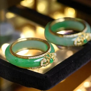 Op de afbeelding: Twee jade armbanden met gouden accenten en diamantversieringen. De jade heeft een levendige groene kleur en de gouden zettingen hebben ingewikkelde ontwerpen. De armbanden worden gepresenteerd op een zwart fluwelen oppervlak, met een vage achtergrond.