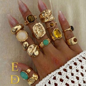 Puede incluir: Una colección de anillos dorados usados en los dedos, con varios diseños que incluyen bandas lisas, superficies texturizadas y engastes con piedras ojo de tigre, blancas, amarillas y turquesas. El texto E D EkmekciDesigns es visible.