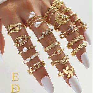 Puede incluir: Una colección de anillos dorados usados en los dedos, con varios diseños que incluyen un motivo de abrazo, perlas, una luna creciente con una estrella, un ojo con una piedra central, una cruz, eslabones de cadena y hojas. El texto E D EkmekciDesigns es visible.