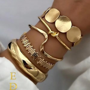 May include: A collection of gold-toned bracelets. The bracelets include a thick, rounded bangle, a textured band, a knot design, and a bracelet with three circular accents. The image also includes the text "E D EkmekciDesigns".