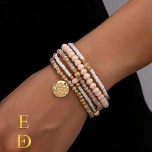 Può includere: Un set di braccialetti di perline bianche, pesca e oro. Un braccialetto presenta un ciondolo a disco dorato. I braccialetti sono indossati su un polso. Il marchio "EkmekciDesigns" è visualizzato in oro.