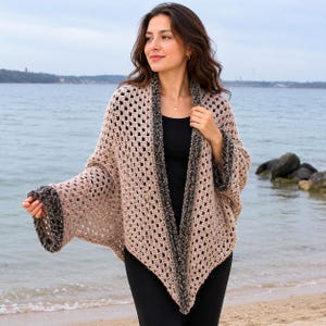 May include: A beige crochet shawl with dark gray trim. The open-weave design features a square pattern. The shawl is worn over a black top. The background shows a beach and water.