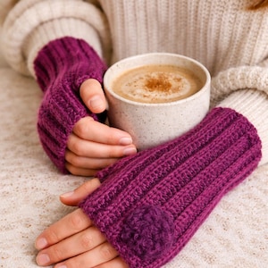 Op de afbeelding: Een persoon draagt een crèmekleurige trui en houdt een mok koffie vast, met een paar paarse vingerloze handschoenen. De handschoenen hebben een geribbeld ontwerp en een kleine decoratieve bloem op elk. De koffie heeft een lichtbruine kleur met een snufje kaneel.
