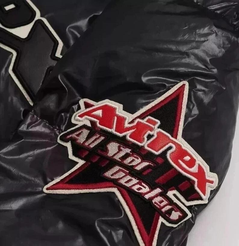 May include: Black puffer jacket with a glossy finish. Features a large patch with the text "Avirex All Star Dealers" in red and white, set against a black and red star design. The jacket has a quilted texture.