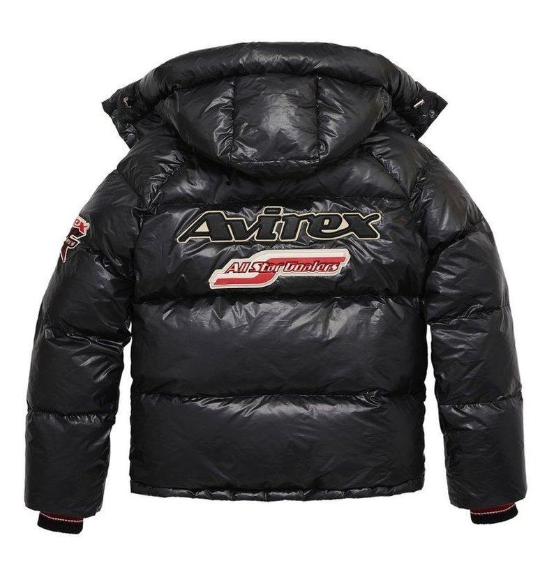 May include: Black puffer jacket with a hood. The jacket features the brand name "Avirex" in gold and white lettering across the back, along with the text "All Star Dunalers" in a red and white design. The jacket has a glossy finish and a red and black striped cuff.