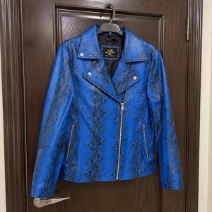May include: A blue faux leather jacket with a snakeskin pattern, hanging on a black hanger. The jacket has a zipper closure, a notched collar, and a small label on the inside. The jacket is a vibrant blue with black detailing.