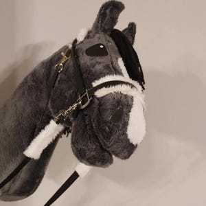 Può includere: Una testa di cavallo a dondolo in peluche grigio con criniera nera e dettagli bianchi. Il cavallo giocattolo presenta una briglia nera con finiture color argento e una redine nera. Il viso del cavallo ha segni bianchi e occhi neri.
