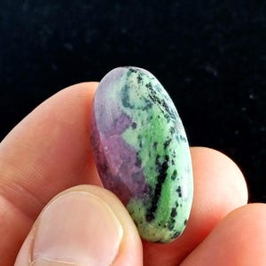 May include: An oval-shaped gemstone with a polished surface. The stone features a mix of colors, including deep red, green, and black. The gemstone is held between fingers, showcasing its size and shape. The overall aesthetic is natural and earthy.